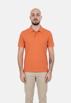 Uomo con testa calva e barba che indossa una polo arancione e pantaloni beige, in piedi con le braccia rilassate davanti a uno sfondo bianco.