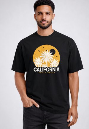 Man with short curly hair and beard wears black T-shirt with white palm trees, orange sun, black moon, and text "CALIFORNIA SOUTH BEACH."