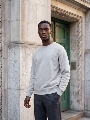 Homme aux cheveux courts portant un sweat-shirt gris clair et un pantalon foncé, debout devant un bâtiment en pierre orné avec une porte verte.