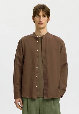 Młody mężczyzna w brązowej koszuli zapinanej na guziki, nałożonej na brązowy t-shirt, oraz zielonych spodniach, stoi na tle jednolitego jasnego tła.