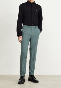 Schwarzes Langarmshirt mit Knopfleistenkragen, kombiniert mit maßgeschneiderten hellgrünen Hosen und schwarzen Anzugschuhen. Logo auf dem Shirt sichtbar.