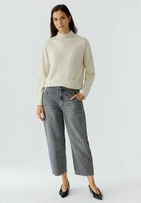 Hellbeiger Pullover mit hohem Kragen und einer Vordertasche, kombiniert mit weiten grauen Jeans. Die Jeans zeichnen sich durch subtile Nähte und einen lockeren Schnitt aus.