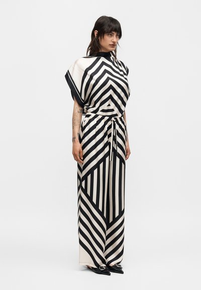 Γυναίκα που φοράει μια ασπρόμαυρη ολόσωμη φόρμα με γεωμετρικές ρίγες, φαρδιά μπατζάκια και κοντά μανίκια, στέκεται μπροστά σε μονόχρωμο φόντο.