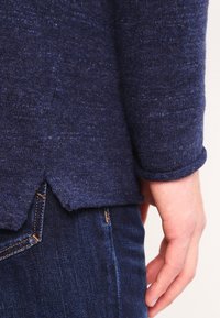 Person som bär en mörkblå stickad tröja med delad nederkant och mörkblå jeans, med handen avslappnad vid sin sida mot en ljus bakgrund.