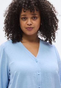 Ανοιχτό μπλε πουκάμισο με υφή, με κουμπιά και βαθύ V λαιμόκοψη και τρία κουμπιά μπροστά. Απαλή υφή, άνετη εφαρμογή και μακριά μανίκια.