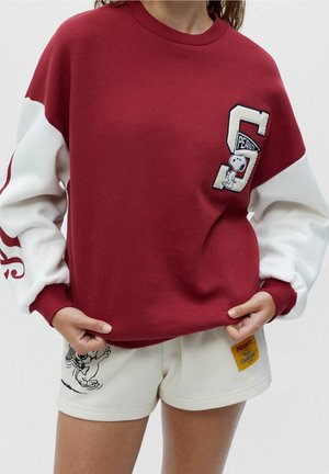 Personne portant un sweat-shirt rouge et blanc avec des patchs Snoopy et Peanuts, assorti à un short blanc avec Woodstock et une étiquette Peanuts.