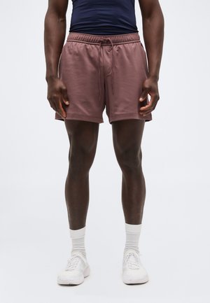 Άνδρας που φοράει λευκά αθλητικά παπούτσια, λευκές κάλτσες και μωβ σορτς με κορδόνι, στέκεται μπροστά σε απλό ανοιχτόχρωμο φόντο.