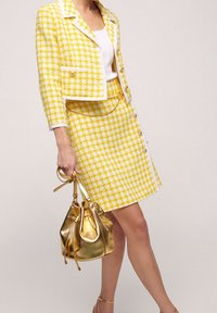 Světlé žluté a bílé kostkované tweedové sako a sukně se zlatými akcenty, doplněné lesklou zlatou kabelkou ve tvaru kbelíku.