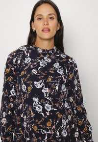 Vestido de manga longa, azul marinho, com padrões florais em branco, rosa e dourado, um colarinho alto e detalhes em botão na parte da frente. Textura de tecido suave.