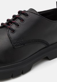 Chaussure en cuir noir avec semelle texturée, lacets rayés noir et rouge, et couture détaillée le long du panneau latéral.