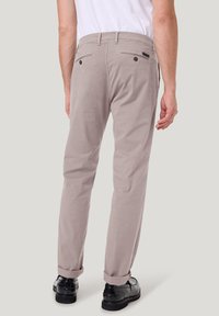 Beige broek met een slim fit, voorzien van opgerolde manchetten, achterzakken met knoopsluitingen en een gladde stofstructuur. Zwarte schoenen zichtbaar.