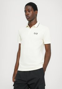 Crème polo shirt met een zwarte kraag en logo op de borst, met korte mouwen en een zachte, gladde textuur. G gedragen met donkere broeken.