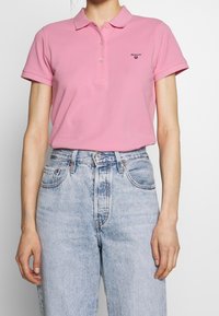 Polo shirt rosa con colletto e tre bottoni, caratterizzato da un piccolo logo sul lato sinistro. Abbinato a jeans a vita alta di colore blu chiaro.