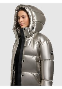 Giacca imbottita color argento metallizzato con ampio cappuccio, cerniera nera e bottoni prominenti. Presenta sezioni trapuntate e una patch con logo sul braccio.