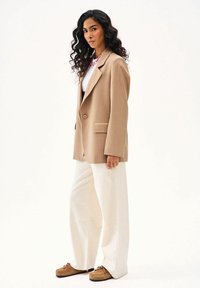 Žena s dlhými kučeravými vlasmi v béžovom nadrozmernom blejzri, bielych nohaviciach a hnedých mokasínach, stojaca na bielom pozadí.