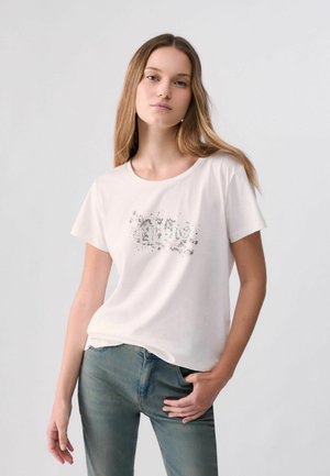 Donna con lunghi capelli castano chiaro indossa una T-shirt bianca con una stampa sbiadita e jeans blu, in piedi con una mano in tasca.