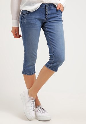Personne portant un pantacourt en jean bleu avec braguette à boutons et des baskets blanches, debout jambes croisées sur fond blanc.