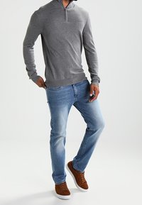 Mann in einem grauen Longsleeve-Zip-Hals-Pullover, blauen Jeans und braunen Wildledersneakern, der vor einem weißen Hintergrund steht.