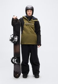 Σνόουμπορντερ που φοράει μαύρο και ελαιπράσινο μονωμένο μπουφάν, μαύρο παντελόνι και κράνος, κρατώντας μια μαύρη σανίδα σνόουμπορντ με λευκές δεστρες.