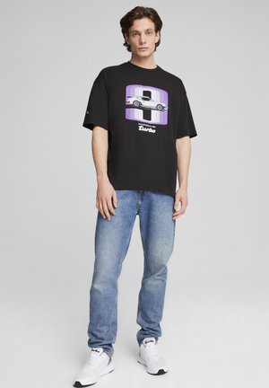 Black oversized t-shirt with a purple graphic design of a car and "Porsche Turbo" text, paired with light blue jeans and white sneakers.