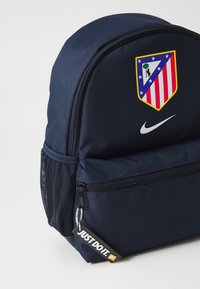 Zaino navy con logo Nike e stemma dell'Atlético Madrid. Presenta un tessuto testurizzato, scomparti con zip e tasca laterale in rete.