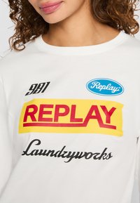 Vrouw in een wit shirt met "Replay," "981," "Replay" in een blauwe ovaal, en "Laundryworks" gedrukt in zwarte cursieve tekst.