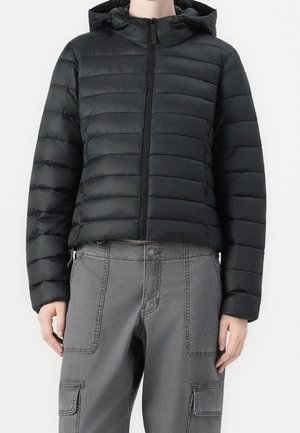 Personne portant une veste matelassée noire à capuche avec une fermeture éclair à l'avant et un pantalon cargo gris avec plusieurs poches.