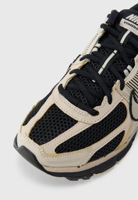 Τα παπούτσια Nike Vomero διαθέτουν μαύρο άνω μέρος από δίχτυ με μπεζ συνθετικ ά επικαλύμματα, μαύρα κορδόνια και υφασμένη μαύρη λαστιχένια σόλα.