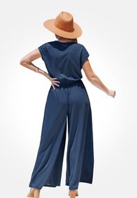 Mono de navy con un corte relajado, mangas cortas, cintura elástica, diseño de pierna ancha, combinado con un sombrero de fieltro marrón y sandalias de tacón.