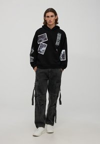 Felpa nera con stampe grafiche bianche; polsini e orlo a coste. Abbinata a pantaloni cargo scuri con più tasche e lacci. Sneakers bianche.