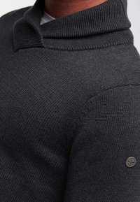 Donkergrijze gebreide trui met een opgevouwen kraag, gestructureerde stof en een klein circulair logodetail op de mouw.