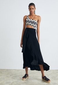Zwarte wikkelskirt met een gerimpelde zoom, gecombineerd met een gemêleerde één-schouder crop top. Het model draagt zwarte sandalen met banden en staat op een gestructureerde vloer.