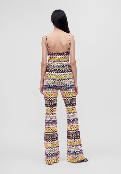 Femme aux longs cheveux foncés se tient de dos, portant un haut sans manches à motif zigzag multicolore et un pantalon évasé assorti.