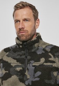 Pullover in fleece con motivo camouflage nei toni di grigio, verde e marrone. Chiusura con zip, colletto alto e leggera texture.