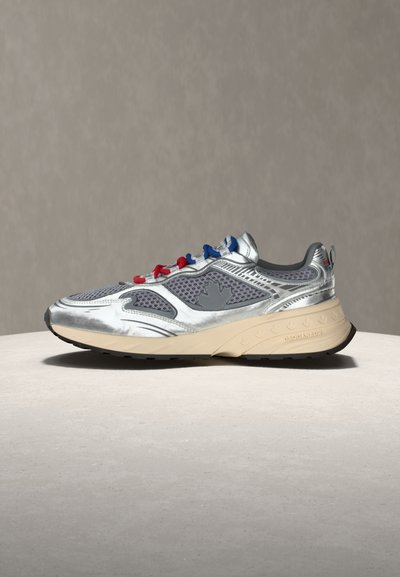 Ασημί και γκρι αθλητικό παπούτσι με κόκκινες και μπλε κορδόνια, μπεζ σόλα και σχέδιο φύλλου σφενδάμου στο πλάι, τοποθετημένο σε ελαφρύ στρογγυλό υπόστρωμα.