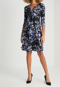 Robe noire à motifs floraux avec un col en V torsadé, des manches trois-quarts et une silhouette fluide, ornée de motifs floraux violets et bleus.