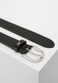 Ceinture en cuir noir avec une texture lisse, boucle en métal argenté et un petit détail de logo près de la boucle. Quatre trous d'ajustement visibles.