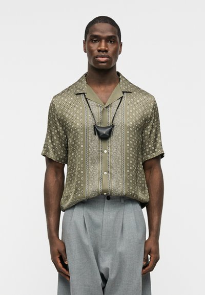 Άνδρας που φοράει κοντομάνικο πουκάμισο με μοτίβο σε ελιά πράσινο χρώμα, γκρι πλισέ παντελόνι και μαύρο κρεμαστό τσαντάκι, στέκεται μπροστά σε απλό φόντο.