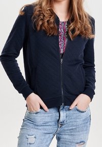 Marinblå quiltad bomberjacka med dragkedja fram, ribbade muddar och elastisk nederkant. Bärs över en mönstrad topp, kombinerad med sliten denim.
