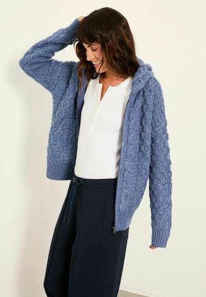 Gestrickte blaue Strickjacke mit einem strukturierten Muster, ausgestattet mit Kapuze, gerippten Bündchen und einem Frontreißverschluss. Kombiniert mit einem weißen Oberteil und dunkler Hose.