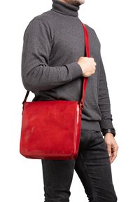 Borsa a tracolla rossa in pelle con design quadrato, texture liscia, cinturino regolabile e una lieve lucentezza, indossata da una persona in un dolcevita grigio.
