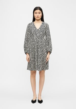 Woman stands facing forward wearing a black and white floral long-sleeve dress with V-neck and black pointed flats on a plain background.