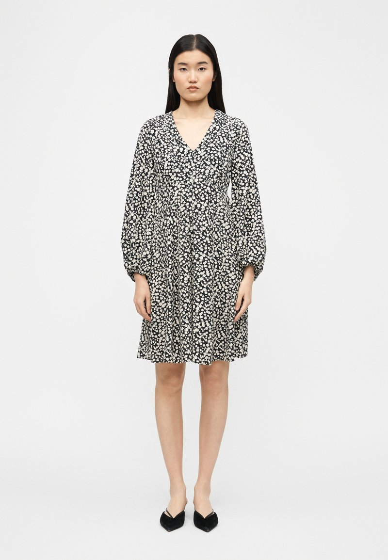 Γυναίκα στέκεται με το πρόσωπο προς τα εμπρός, φορώντας μαύρο-άσπρο μακρυμάνικο φόρεμα με λουλουδένιο σχέδιο και V-λαιμόκοψη, καθώς και μαύρα μυτερά παπούτσια σε απλό φόντο.