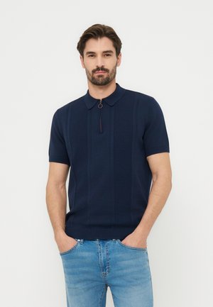 Bărbat cu păr scurt și întunecat și barbă, purtând un polo tricotat cu mâneci scurte, culoare bleumarin, cu fermoar în față, și blugi albaștri, stând cu mâinile în buzunare.