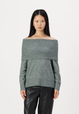 Pull doux gris clair, à épaules nues, avec une texture côtelée, coupe ample et décolleté large. Associé à un pantalon en cuir noir brillant.