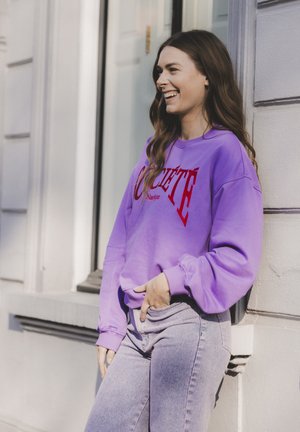 Paarse sweatshirt met rode tekst, gecombineerd met lichtblauwe jeans. De stof lijkt zacht met een comfortabele pasvorm en geribbelde manchetten.