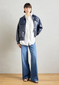 Jūras zilas spīdīgas mākslīgās ādas stiprinājuma jakas, baltas pogkrekli, gaiši zilas izpleša džinsus un melnas sandaļas. Modelis stāv uz koka grīdas.