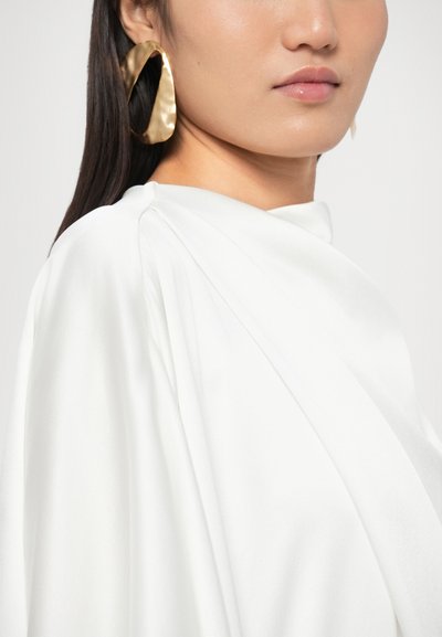 Bijeli svileni top s naboranim dizajnom i mekom teksturom, uparen s velikim, kovanim zlatnim naušnicama u obliku obruča koje reflektiraju svjetlost.