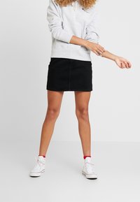Schwarzer Jeansrock mit taillierter Form, kombiniert mit einem hellgrauen Kapuzenpullover und weißen High-Top-Sneakern mit roten Akzenten.
