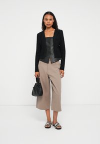 Kvinde iført en sort tekstureret cardigan, sort knaplædt læderkorsæt, beige brede cropped bukser, sandaler med slangeprint, mens hun holder en sort håndtaske.